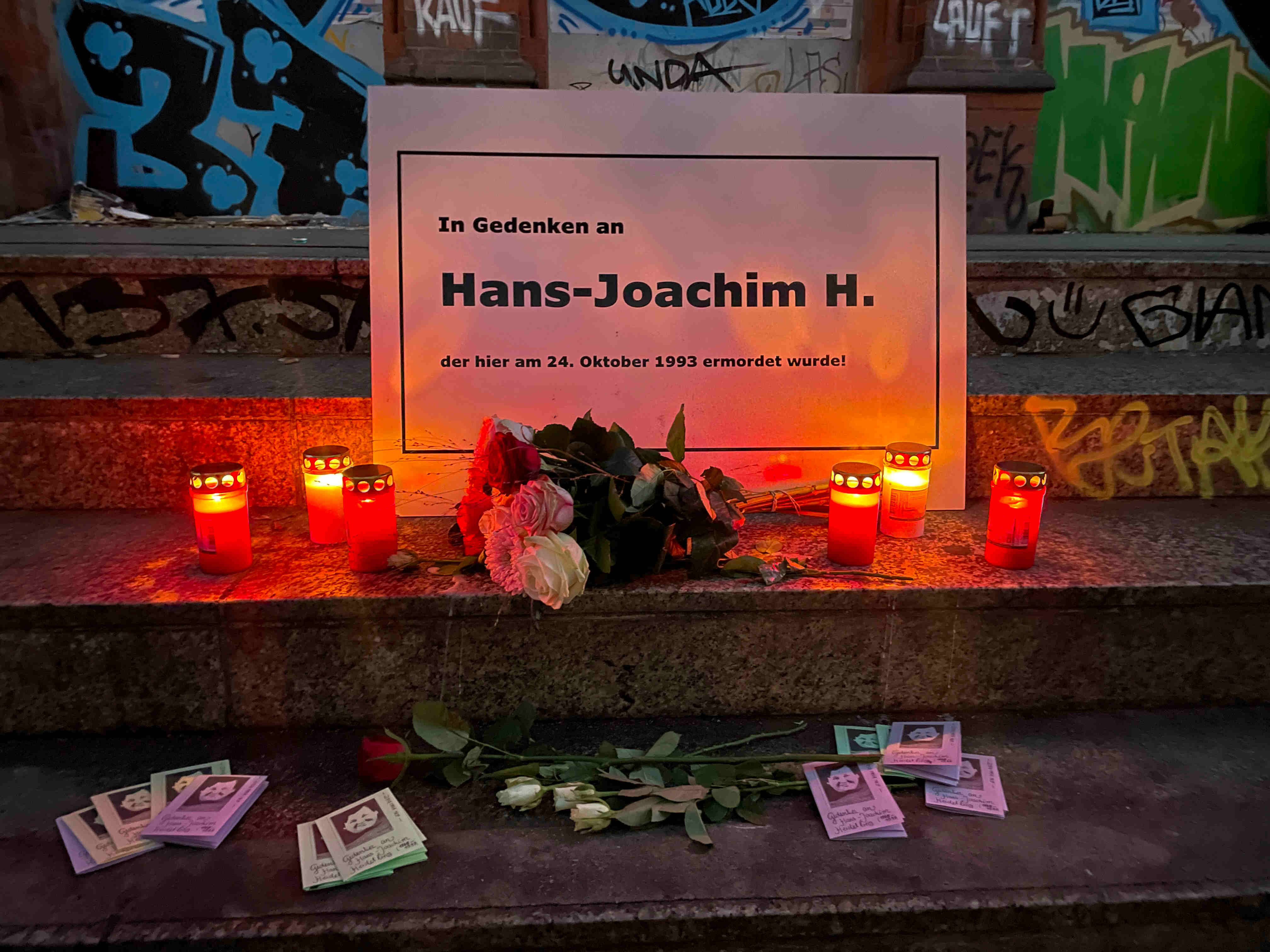 Foto vom Gedenken vor dem S-Bahnhof-Schöneweide. Auf der Treppe steht eine Gedenktafle mit den Worten '"In Gedenken an Hans-Joachim H. der hier am24. Oktober 1993 ermordet wurde!", davor liegt ein Strauß Rosen, sowie 6 Grablichter rechts und links daneben, darunter liegen Mini-Zines mit Informationen über Hans-Joachim Heidelberg und der Gedenkinitiative.